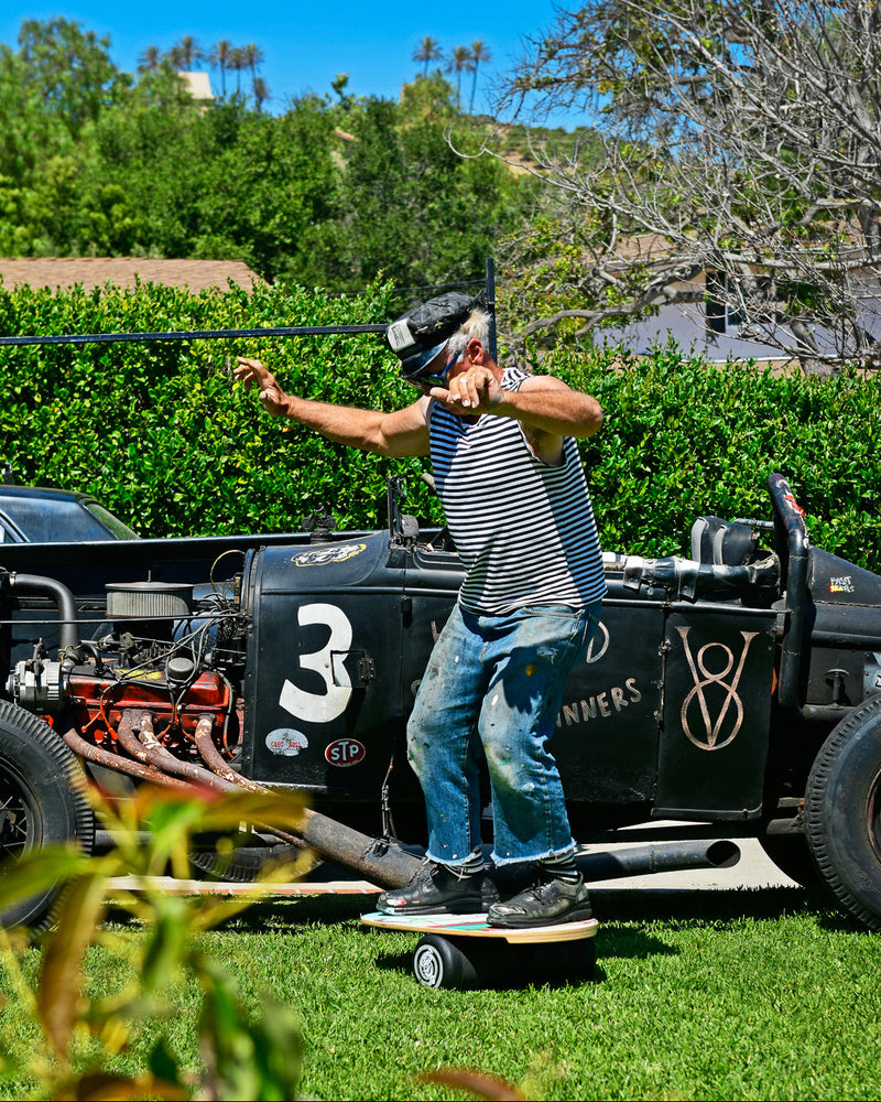 Indo Board balance trainer in use outdoors by a man wearing a VR headset and striped tank top, positioned near a vintage car.